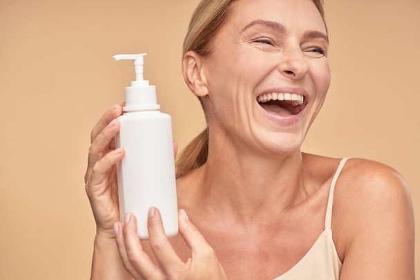 Cheerful adult lady holding cosmetic cream while standing in studio - Photo, Image