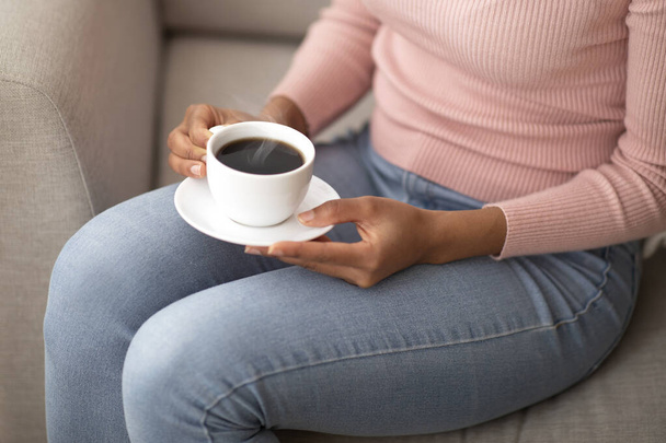 Przerwa na kawę. Widok upraw czarnej kobiety trzymając filiżankę aromatycznego napoju podczas siedzenia na kanapie w pomieszczeniach, zbliżenie - Zdjęcie, obraz