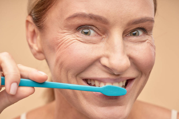 Hammashygienia. Hymyilevä kypsä nainen huolehtii suuontelosta sinistä hammasharjaa käytettäessä - Valokuva, kuva