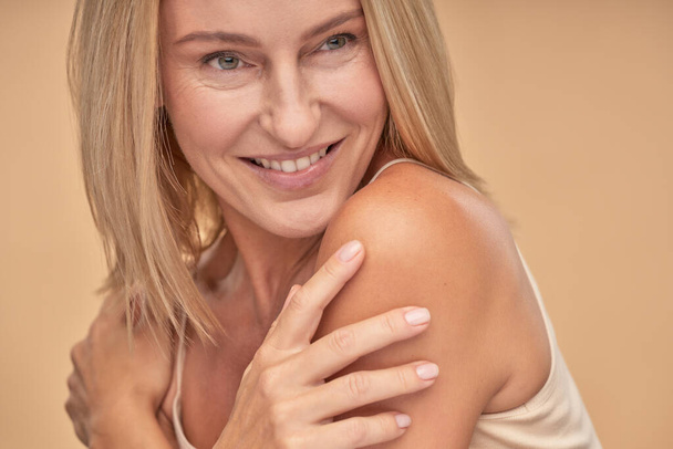 Portrait of beautiful adult woman touching her shoulders with her hands and looking aside while posing in studio over beige background - Photo, Image