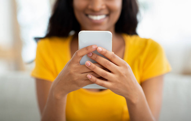 Oříznutý pohled na pozitivní černošku držící moderní smartphone uvnitř, chatující s přáteli nebo posílající email - Fotografie, Obrázek