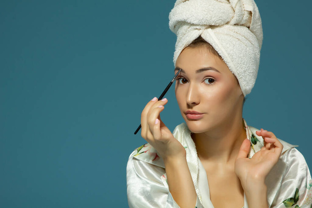 Young attractive woman putting tone on her eyebrows over blue background. Charming girl makes morning beauty treatments and make-up - Zdjęcie, obraz