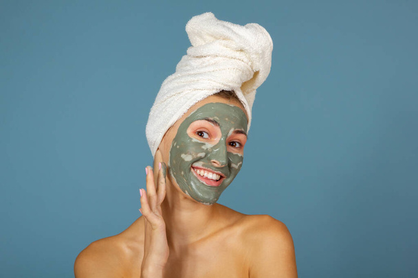 Beautiful cheerful teen girl applying facial clay mask. Beauty treatments, isolated over blue background. - Фото, зображення