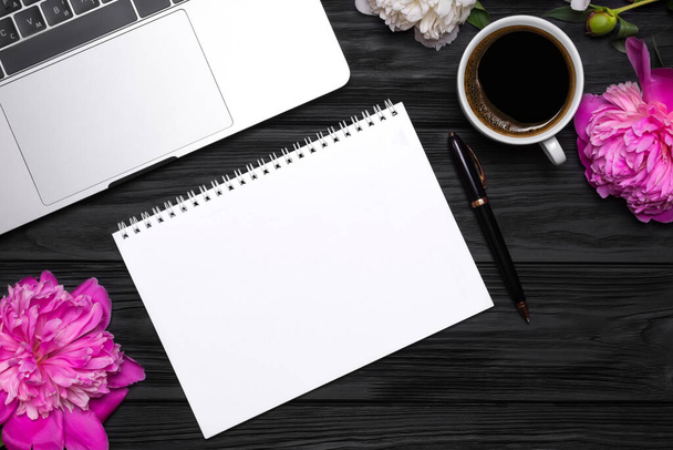 Παιώνια λουλούδια, καφέ, στυλό, laptop και άδειο σημειωματάριο για το κείμενό σας σε μαύρο ξύλινο φόντο. - Φωτογραφία, εικόνα