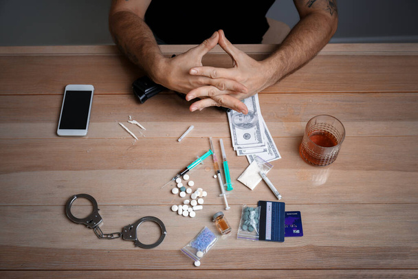 Мужчина-наркоман с наркотиками, деньгами, алкоголем и наручниками сидит за столом, вид сверху. Концепция зависимости - Фото, изображение