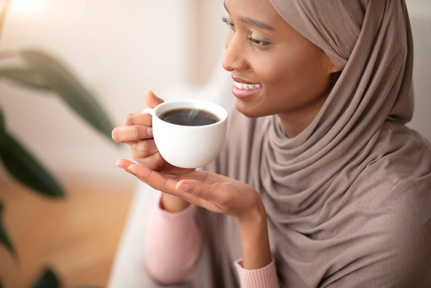 Szczęśliwy poranek. Szczęśliwa muzułmanka w tradycyjnym hidżabie pijąca mocną czarną kawę w domu - Zdjęcie, obraz