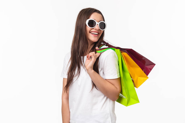 Shopping, leisure and lifestyle concept. Happy, joyful young feminine woman walking in store, buying new clothes and summer bikini, look back with smiling pleased look, hold bags on shoulder - Photo, Image