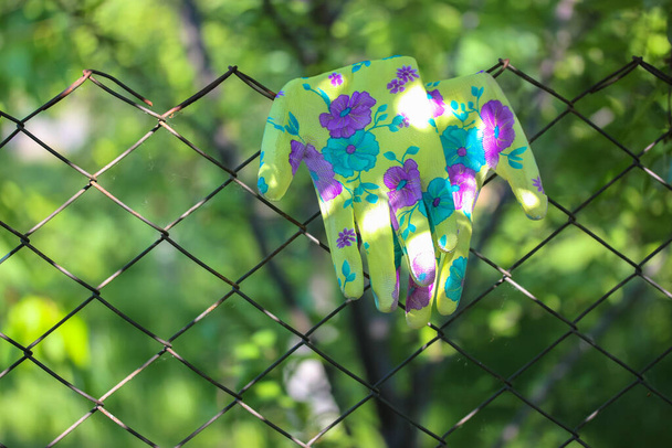 Romper en el jardín en verano, guantes verdes de colores para trabajar en el jardín en una valla de metal en un árbol de fondo borroso - Foto, Imagen