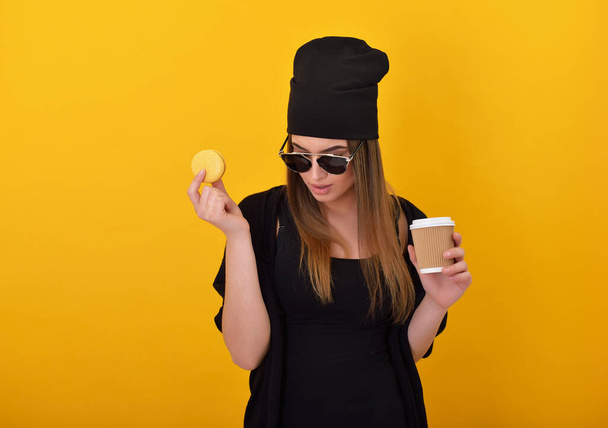 Hipster girl eats cake macaron and drinks cogffee over yellow background. Studio female portrait with sweet food and hot drink. - Photo, image
