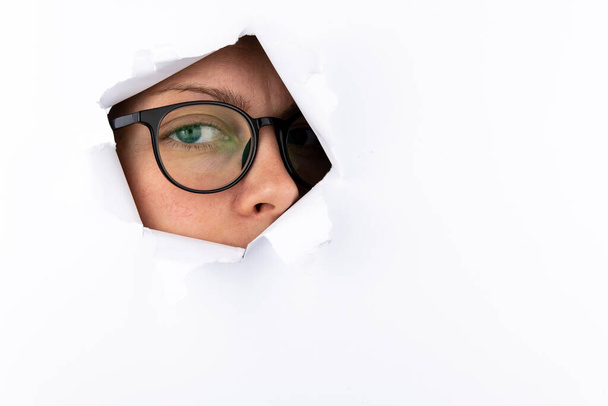 A woman's eye with black-framed glasses looks through a hole in a white paper wall. Espionage concept. Free space for text. - Fotoğraf, Görsel