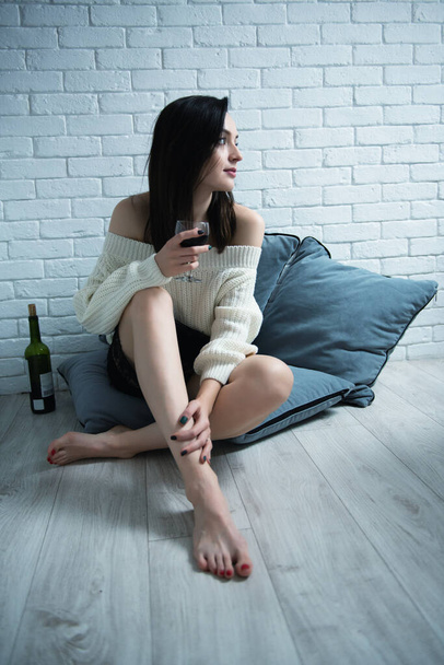 Portrait of charming young woman sitting on the floor at home and drinking red wine, image toned and noise added - Foto, Imagem