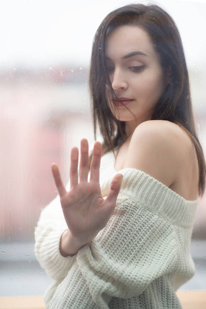 Young beautiful woman with long dark hair dressed in a white sweater, outdoor behind window pane on which are raindrops.  Lady, portrait at home. Self isolation, stay at home, quarantine concept - Photo, image