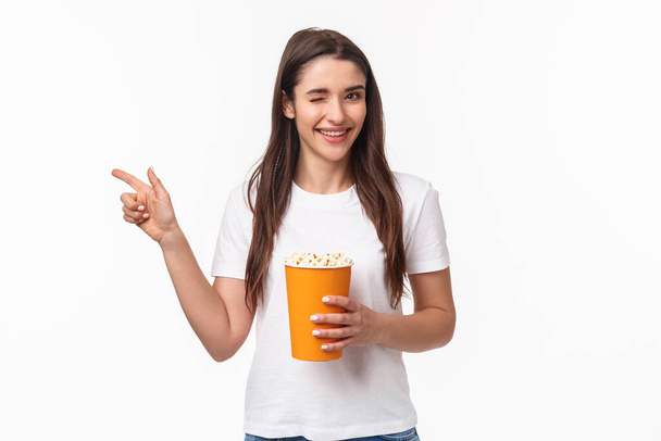Entertainment, fun and holidays concept. Portrait of cheeky cute young girl in t-shirt, wink and smiling, pointing finger left inviting come and watch movie together in cinema, hold popcorn - Photo, Image