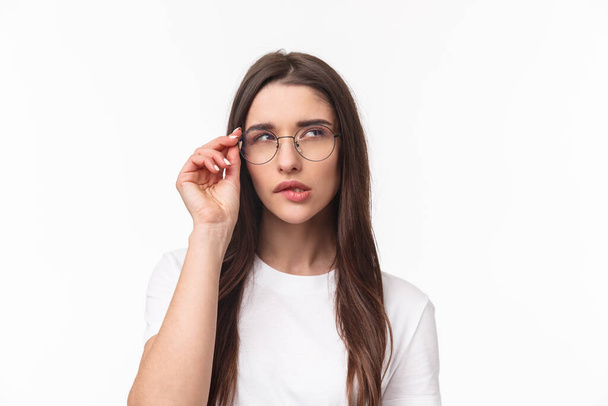 Close-up portrait of smart and thoughtful beautiful young girl in glasses, thinking, squinting suspicious and biting lip look up while pondering something, have interesting idea, white background - Photo, Image