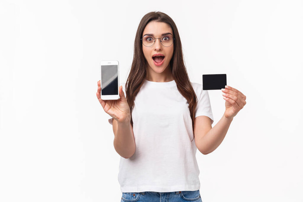 Портрет взволнованной и счастливой молодой женщины представить новое приложение, любимый интернет-магазин, чтобы купить одежду, показать мобильный телефон и кредитную карту, улыбаясь весело, белый фон - Фото, изображение