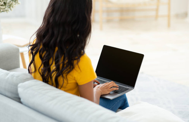 Неузнаваемая черная леди, печатающая на ноутбуке с чистым экраном на удобном диване дома, макет для дизайна - Фото, изображение