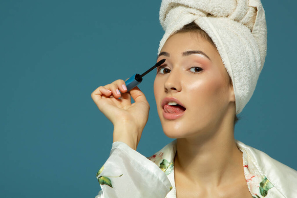 Young attractive funny woman putting eye mascara, studio shot over blue background, beauty treatment morning  - 写真・画像