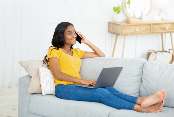 Fröhliche Afroamerikanerin kommuniziert von zu Hause aus per Handy und Laptop mit jemandem - Foto, Bild