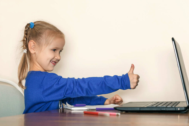 Klein meisje in een blauwe blouse stak haar vinger op. Het kind zit voor de laptop en wijst naar de monitor duimen omhoog - Foto, afbeelding