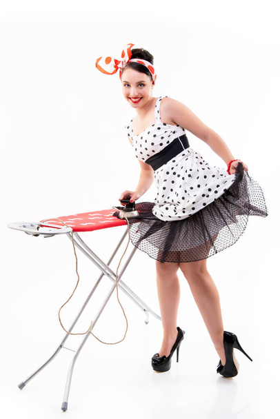 Pinup girl irons her dotted dress and happy smiling. Retro portrait of young cheerful woman in pin-up style, vintage stylization, studio shot over white background.  - 写真・画像