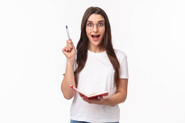 Portrait of enthusiastic, creative young woman in glasses, t-shirt, raising pen up eureka gesture, have moment of realisation, got an idea, holding planner or diary, writing ideas in notebook - Фото, изображение