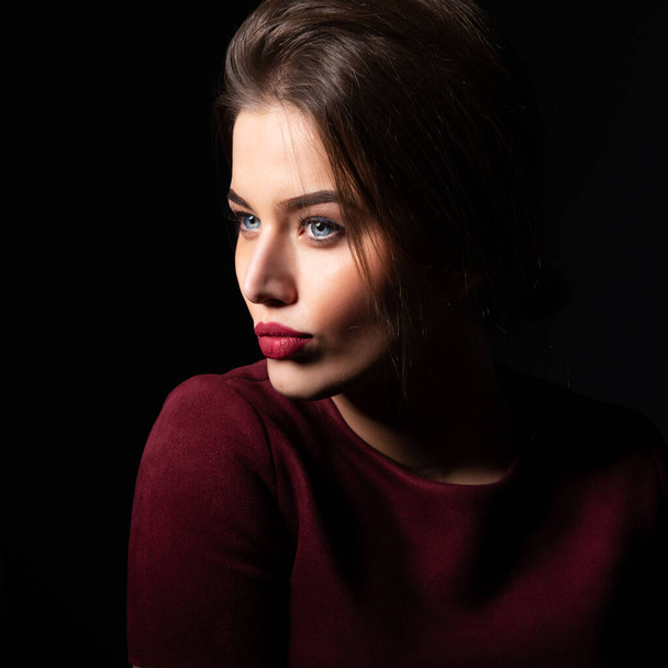 Charming young woman with blue eyes and long fair hair posing at studio over dark background. Amazing stylish girl with perfect makeup wearing elegant deep red dress. Beauty and fashion - Foto, imagen