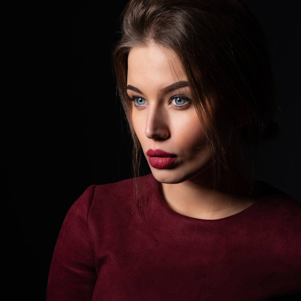 Charming young woman with blue eyes and long fair hair posing at studio over dark background. Amazing stylish girl with perfect makeup wearing elegant deep red dress. Beauty and fashion - Photo, image