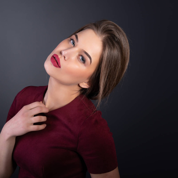 Charming young woman with blue eyes and long fair hair posing at studio over dark background. Amazing stylish girl with perfect makeup wearing elegant deep red dress. Beauty and fashion - Fotó, kép