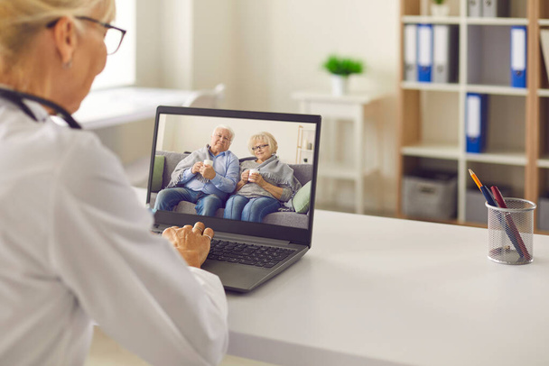 Doctor sitting at a table in front of a laptop and talking to an elderly couple via video link. - Foto, Imagem