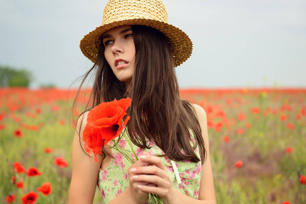 Young beautiful woman wearing straw hat and a green dress walking at poppy field. Happy girl dancing among blooming red flowers, spring nature outdoor  - Valokuva, kuva
