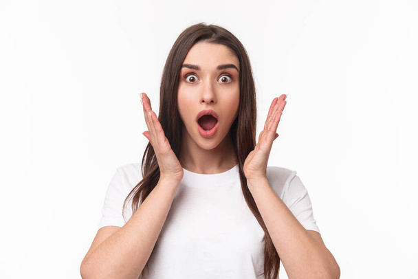 What an amazing news. Surprised and astonished, excited young woman react to something awesome happened, gasping, open mouth and raise hands near face, staring camera, white background - Foto, Imagem