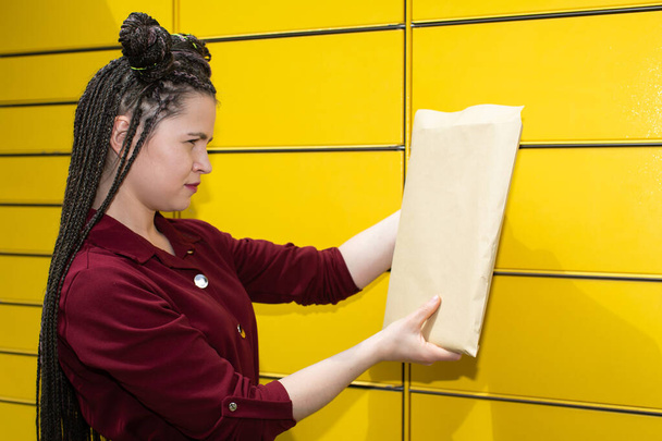 A young adult prepares a shipment in a gray envelope to be sent through a parcel machine. African braids. - Photo, Image