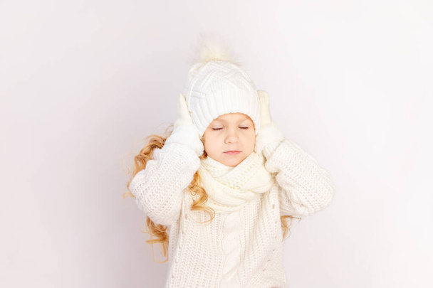 child girl in a sweater and hat holding her head with closed eyes on a white isolated background, space for text - Foto, imagen