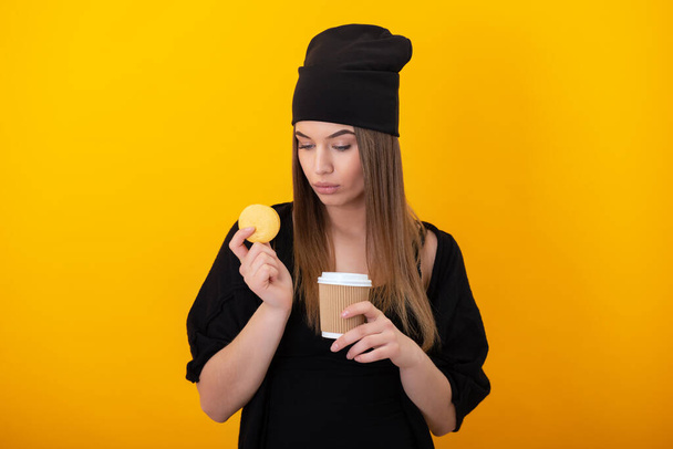Stylish girl eats cake macaron over yellow background. Studio female portrait with sweet food and drink ,take and go. Woman drinks coffee and eats dessert - Photo, image