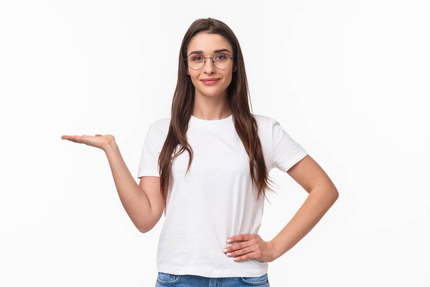 Waist-up portrait of pleasant good-looking caucasian female in glasses, attractive girl holding someone on hand, raising arm as have product on it, smiling camera, advertise, white background - Photo, Image