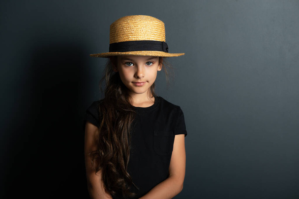 Beautiful caucasian little girl wearing black t0shirt and straw hat posing over deep gray wall looking at camera. Fashion portrait of cute kid.  - Foto, Imagem
