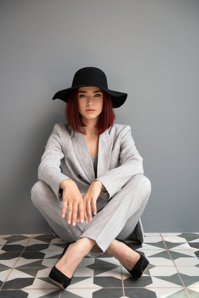 Beautiful fashion woman. Full length portrait of beautiful girl wearing light gray suit and black hat posing indoor over deep  gray wall - Фото, зображення
