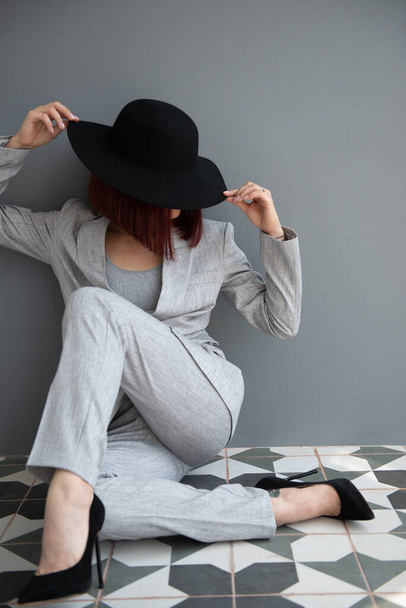 Beautiful fashion woman. Full length portrait of beautiful girl wearing light gray suit and black hat posing indoor over deep  gray wall - Фото, зображення