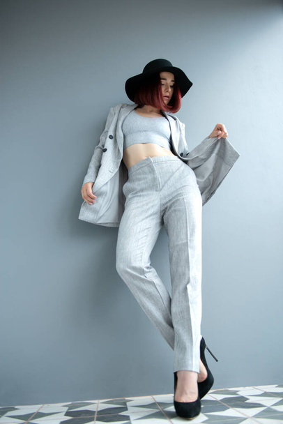 Beautiful fashion woman. Full length portrait of beautiful girl wearing light gray suit and black hat posing indoor over deep  gray wall - Zdjęcie, obraz