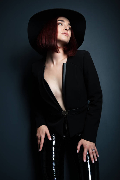 Beautiful fashion woman with perfect body. Portrait of beautiful girl wearing black blazer, latex tight pants and hat posing in studio over deep  gray background - Foto, Imagem