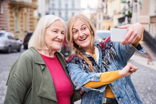 Woman inviting her senior stylish mom to take a selfie - Foto, afbeelding