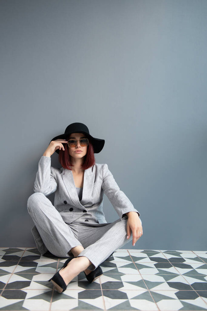 Beautiful fashion woman. Full length portrait of beautiful girl wearing light gray suit and black hat posing indoor over deep  gray wall - Foto, Imagen