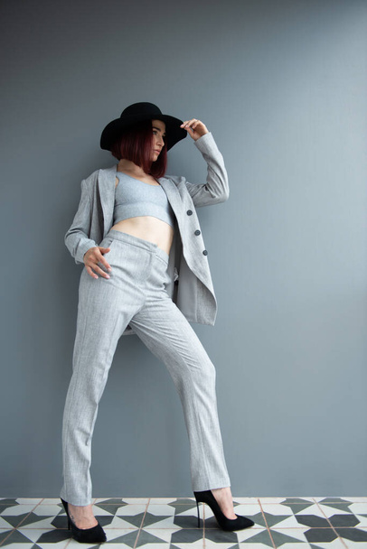 Beautiful fashion woman. Full length portrait of beautiful girl wearing light gray suit and black hat posing indoor over deep  gray wall - 写真・画像