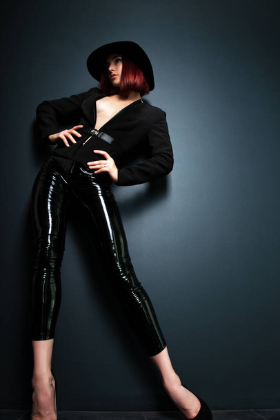 Beautiful fashion woman with perfect body. Portrait of beautiful girl wearing black blazer, latex tight pants and hat posing in studio over deep  gray background - 写真・画像