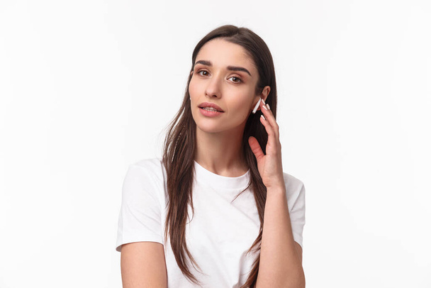 Lähikuva muotokuva naisellinen houkutteleva nuori nainen t-paita, koskettaa langatonta nappikuulokkeet, kuunnella musiikkia kuulokkeet, näköinen kamera, päälle podcast tai rentouttava soittolista, valkoinen tausta - Valokuva, kuva