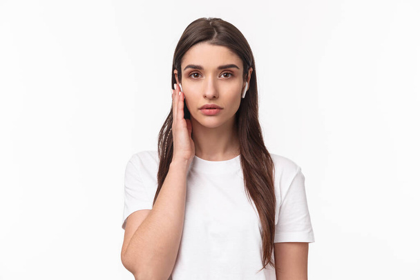 Close-up portrait of serious-looking young confident female asnwering calls using wireless headphones, touching earbud to change volume or answer call, look camera determined, white background - Foto, Imagem