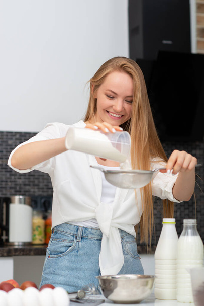 Young woman standing in the kitchen at home and cooking with enjoyment bakery products of flour, milk, cocoa, sugar and fruits. Funny portrait of happy girl chef sifts flour - Foto, afbeelding
