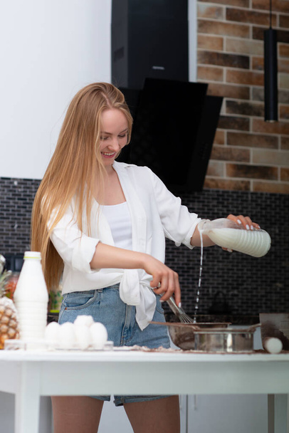 Young woman standing in the kitchen at home and cooking with enjoyment bakery products of flour, milk, cocoa, sugar and eggs. Funny portrait of happy girl chef  - 写真・画像