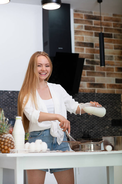 Young woman standing in the kitchen at home and cooking with enjoyment bakery products of flour, milk, cocoa, sugar and eggs. Funny portrait of happy girl chef  - Fotó, kép