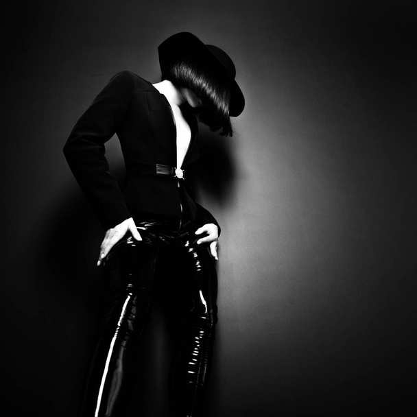 Beautiful fashion woman with perfect body. Portrait of beautiful girl wearing black blazer, latex tight pants and hat posing in studio over deep  gray background, black and white - 写真・画像
